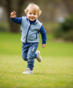 Photograph-of-a-happy-blonde-toddler-in-blue-vest-and-jeans-running-on-grassy-field-with-blurred-trees-in-background