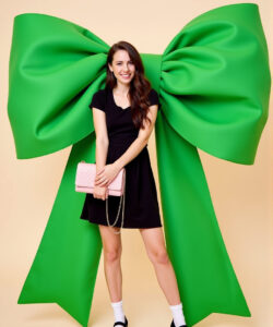 Photograph-of-a-smiling-young-woman-in-a-black-dress-holding-a-pink-Chanel-handbag-against-a-giant-green-bow