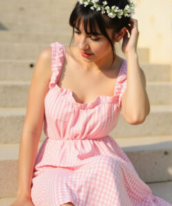 Photograph-of-an-Asian-woman-in-pink-gingham-dress-white-flower-crown-sitting-on-stone-steps-soft-sunlight