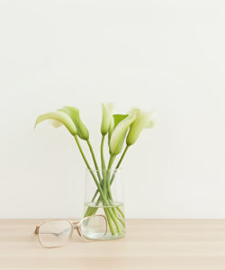Photograph-of-pale-green-calla-lilies-in-a-glass-vase-with-a-pair-of-gold-framed-glasses-on-a-light-wooden-surface
