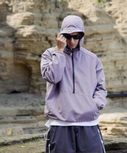 The-photograph-captures-a-young-man-standing-in-a-rugged-outdoor-setting-with-a-backdrop-of-beige-rocky-cliffs-and-a-shallow-stream-He-is-wearing-a-light-lavender-hooded-sweatshirt-with-a-zipper-front