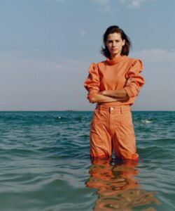 The-photograph-features-a-young-woman-standing-in-shallow-water-with-the-ocean-as-the-background-She-has-a-slender-build-and-is-wearing-a-bright-orange-jumpsuit-with-long-sleeves-and-a-high-collar-which