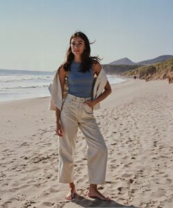 This-is-a-high-resolution-photograph-of-a-young-woman-standing-on-a-sandy-beach-with-a-clear-blue-sky-and-gentle-waves-in-the-background-The-woman-is-positioned-slightly-to-the-left-of-the-center-facing