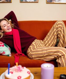 This-is-a-photograph-of-a-young-woman-lounging-on-a-burnt-orange-sofa-in-a-cozy-living-room-She-has-light-blonde-hair-and-is-wearing-a-maroon-sweater-and-a-bright-red-scarf-paired