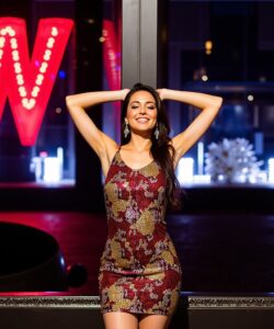 This-is-a-photograph-of-a-young-woman-standing-in-front-of-a-modern-building-with-a-large-illuminated-sign-reading-W-in-bright-red-The-woman-is-of-medium-height-and-slender-build-with