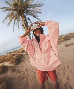 This-is-a-photograph-of-a-young-woman-standing-on-a-sandy-beach-with-a-clear-blue-sky-and-a-palm-tree-in-the-background-She-is-wearing-a-large-light-pink-puffer-jacket-with-a-hood
