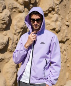 a-young-man-standing-in-a-desert-like-setting-with-beige-rock-formations-in-the-background-The-man-is-wearing-a-light-lavender-windbreaker-jacket-with-a-hood