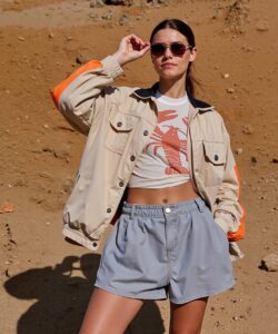 a-young-woman-standing-against-a-rugged-sandy-background-The-woman-is-positioned-slightly-off-center-facing-the-camera-with-her-right-hand-holding-her-sunglasses-which-are-perched-on