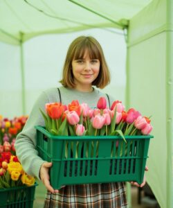 a-young-woman-standing-under-a-light-green-canopy-holding-a-large-green-plastic-basket-filled-with-vibrant-tulips-She-has-shoulder-length-straight-brown-hair-with-bangs-and-a-light