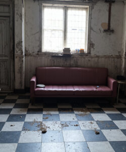 A-dilapidated-abandoned-room-with-a-red-couch-checkered-floor-wooden-cabinets-and-a-window-devoid-of-people