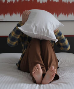 A-person-sits-on-a-bed-hugging-a-pillow-with-feet-up-wearing-a-plaid-shirt-and-brown-pants