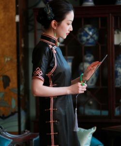 A photograph of a slender asian woman in traditional black cheongsam with red trim holding a fan in an elegant room