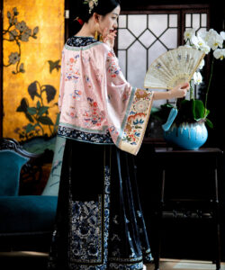 A photograph of an elegant chinese woman in traditional pink and black cheongsam holding a fan in a luxurious room
