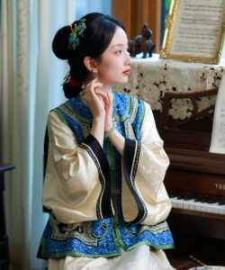 A photograph of an elegant east asian woman in traditional blue and gold attire seated by a piano indoors