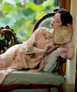 A serene asian woman in traditional pink floral robe reclines on vintage armchair holding a fan by a window
