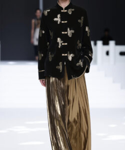 Asian model in black velvet jacket with gold butterflies gold pleated skirt walking on white runway minimalistic background