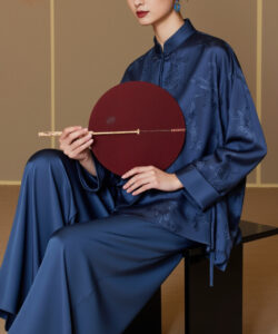 Asian woman in blue silk robe and pants holding a red fan sitting on a bench elegant and serene
