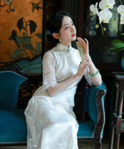 Asian woman in white lace dress holding fan sitting on blue velvet sofa elegant room with floral wallpaper orchid