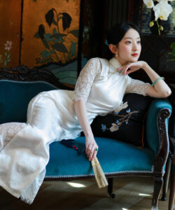 Asian woman sitting on a vintage blue velvet sofa wearing a white lace dress holding a fan elegant setting
