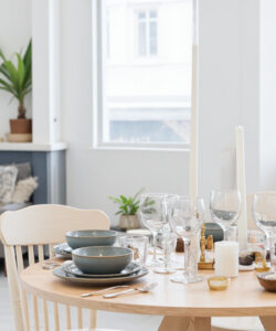 Bright-modern-dining-room-with-light-wooden-table-grey-plates-white-chairs-and-potted-plants-by-large-windows