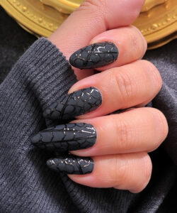 Close up photograph of a hand with black crackled nail polish wearing a grey sweater against a dark background
