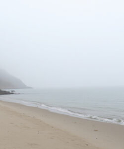 Foggy-coastal-scene-with-lighthouse-on-left-sandy-beach-calm-sea-and-distant-ship-on-right