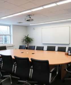 Modern-spacious-conference-room-with-a-large-wooden-oval-table-black-chairs-white-walls-large-windows-and-a-projector-screen