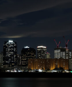 Nighttime-photograph-of-a-city-skyline-with-illuminated-skyscrapers-cranes-and-a-river-in-the-foreground
