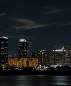 Nighttime-photograph-of-a-city-skyline-with-lit-skyscrapers-cranes-and-a-calm-river-reflecting-city-lights
