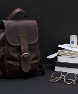 Photograph-Brown-leather-backpack-glasses-books-and-a-jar-of-honey-on-a-dark-surface