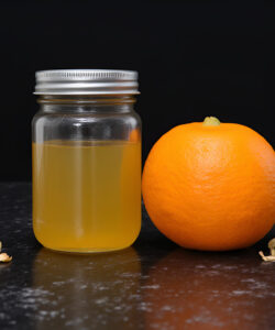 Photograph-Clear-glass-jar-with-yellow-liquid-whole-orange-scattered-oat-flakes-small-yellow-flowers-on-black-marble-surface