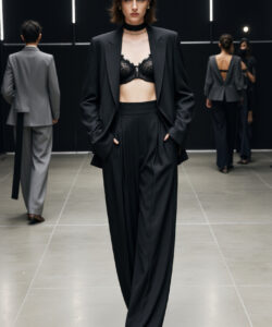Photograph dark haired woman in black suit lace bra choker standing confidently on a minimalist dimly lit runway with other models