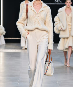 Photograph elegant white haired model in cream knit sweater and pants holding beige handbag struts on runway amidst fur clad models
