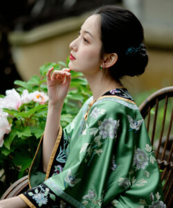 Photograph of a chinese woman in a green floral kimono sitting outdoors holding a flower with potted plants nearby