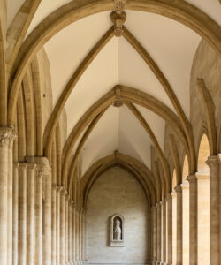 Photograph-of-a-Gothic-style-cloister-with-tall-arched-columns-stone-walls-and-a-small-statue-in-the-central-alcove