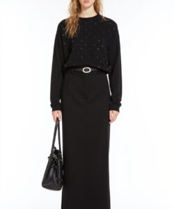 Photograph of a blonde woman in black sweater high waisted skirt black boots and handbag standing against a plain white background