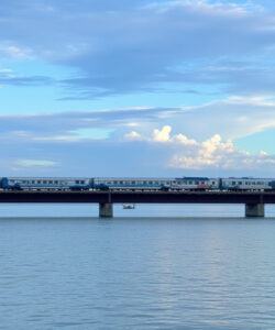 Photograph-of-a-blue-and-white-train-crossing-a-bridge-over-calm-waters-under-a-partly-cloudy-sky
