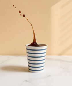 Photograph-of-a-blue-striped-ceramic-cup-spilling-dark-liquid-onto-a-white-marble-surface-against-a-beige-wall