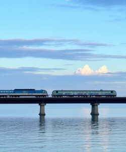 Photograph-of-a-blue-train-on-a-bridge-over-calm-water-with-a-sky-filled-with-soft-clouds