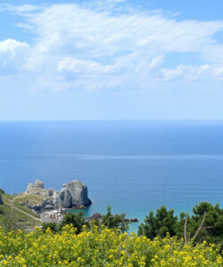 Photograph-of-a-coastal-landscape-with-a-small-red-roofed-stone-building-rocky-cliffs-a-distant-island-and-a-vibrant-blue-sea