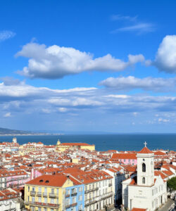Photograph-of-a-coastal-town-with-red-roofed-buildings-clear-blue-sky-scattered-white-clouds-and-distant-mountains