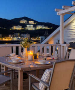 Photograph-of-a-cozy-outdoor-dining-setup-on-a-wooden-deck-at-dusk-overlooking-a-lit-coastal-town