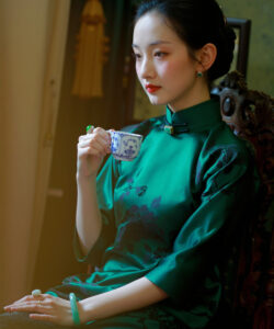 Photograph of a delicate asian woman in a green cheongsam holding a blue floral teacup with a serene expression