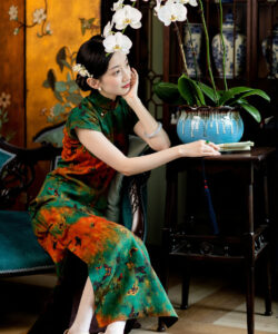 Photograph of a delicate asian woman in traditional green and orange qipao seated by a blue vase with white orchids