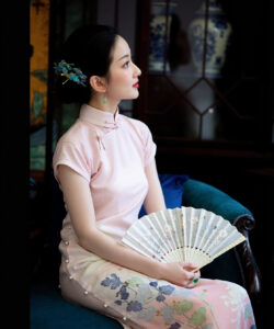 Photograph of a fair skinned asian woman in a pink cheongsam holding a floral fan seated on a blue velvet chair