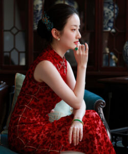 Photograph of a fair skinned asian woman in a red cheongsam seated in a dimly lit room with antique decor