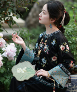 Photograph of a fair skinned asian woman in traditional black floral silk robe holding a green fan amidst blooming pink flowers