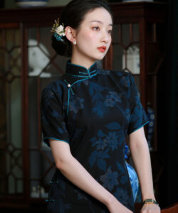 Photograph of a fair skinned dark haired asian woman in a black floral cheongsam standing in a room with blue porcelain