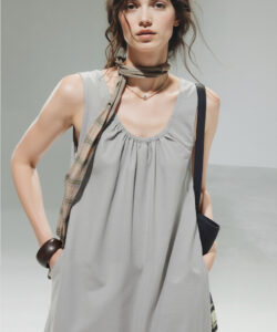 Photograph of a fair skinned dark haired woman in a sleeveless gray dress plaid scarf and black handbag set against a gradient