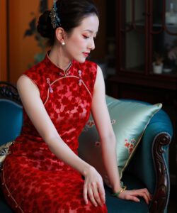 Photograph of a fair skinned woman in a red floral cheongsam seated on a blue sofa with ornate background
